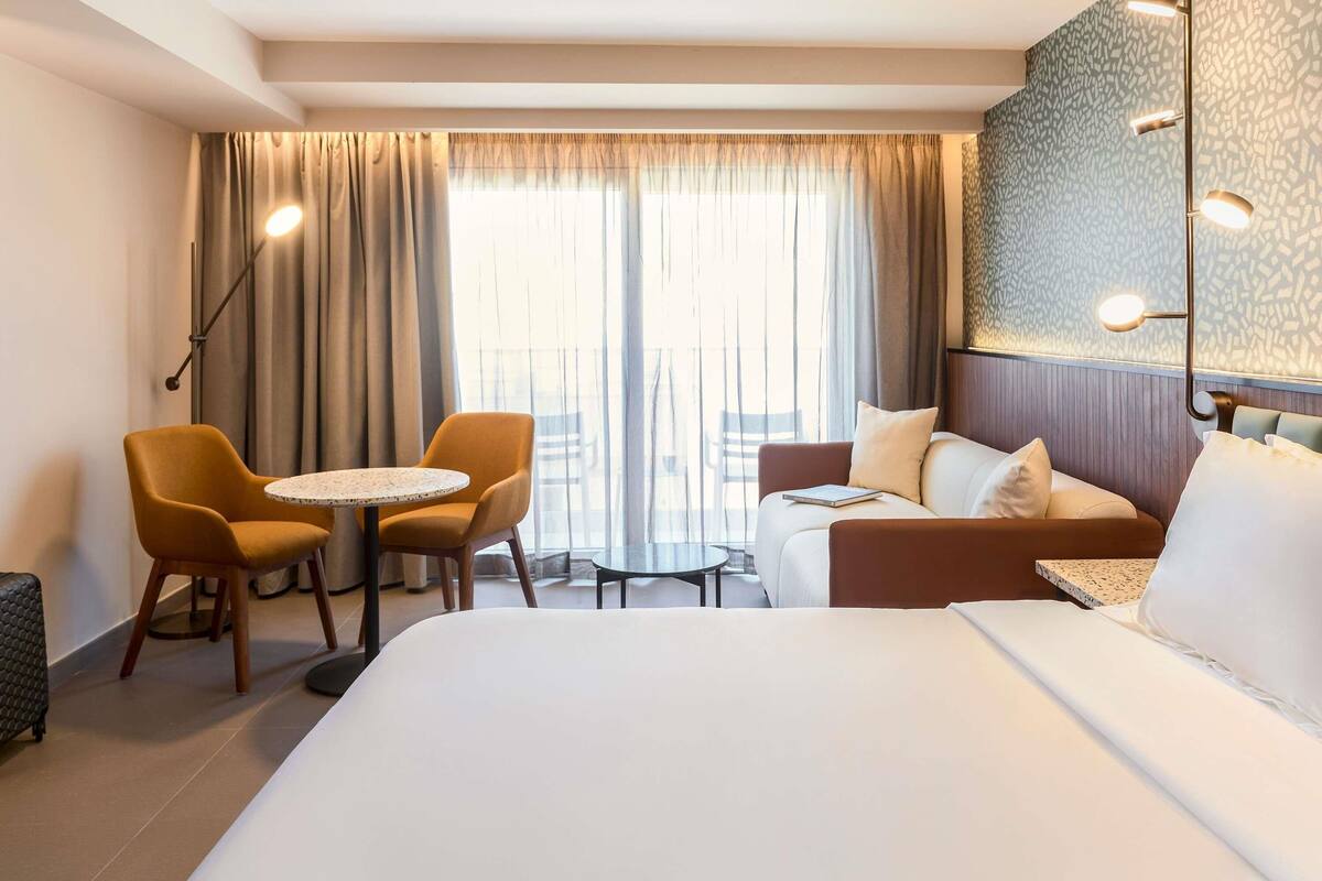 A hotel room with a large bed, a sofa, two armchairs, a small round table, and a large window with curtains.