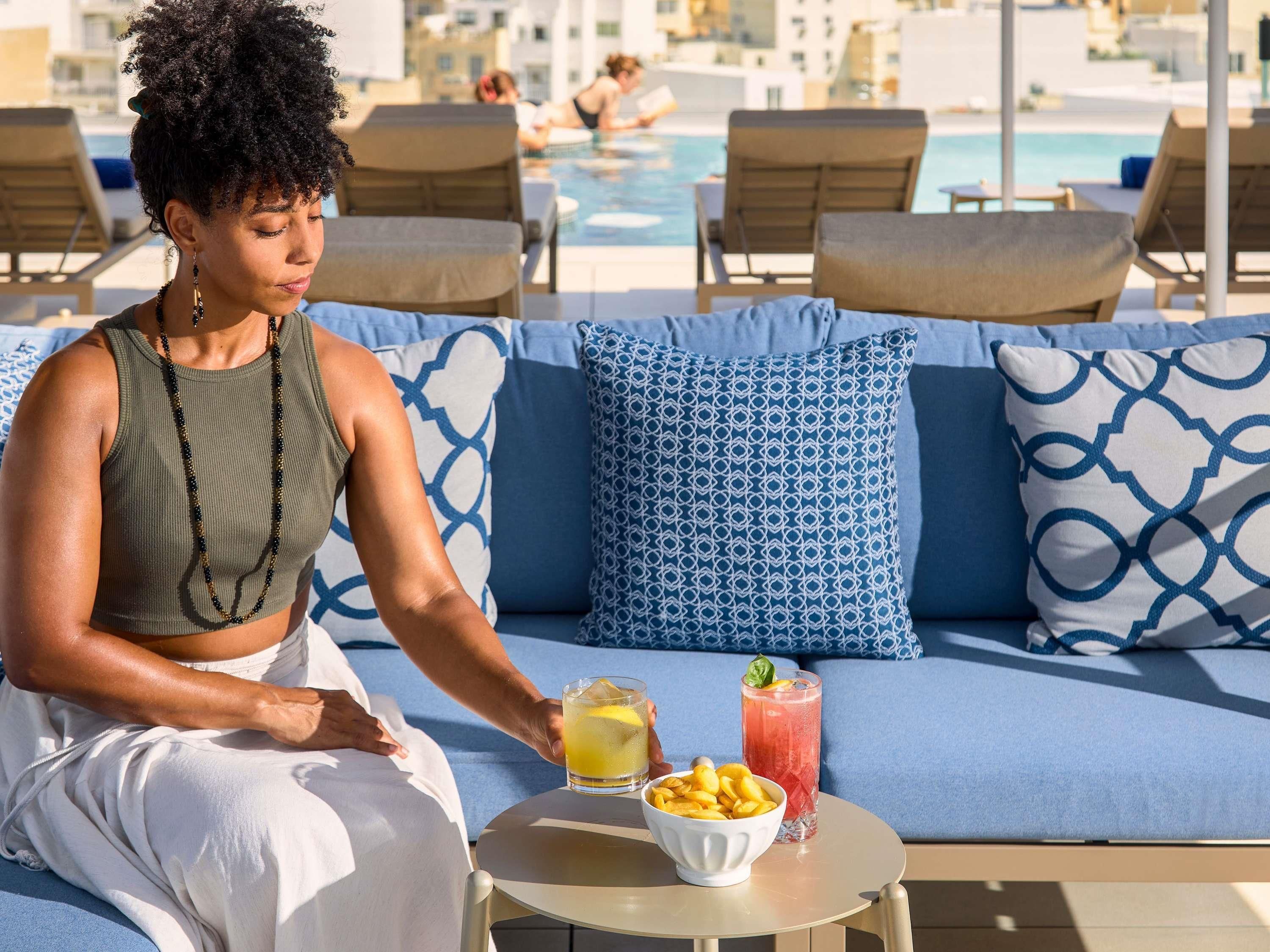 Une personne est assise sur un canapé bleu agrémenté de coussins à motifs. Devant elle, une petite table ronde porte une boisson et quelques en-cas. On aperçoit une piscine à l’arrière-plan.