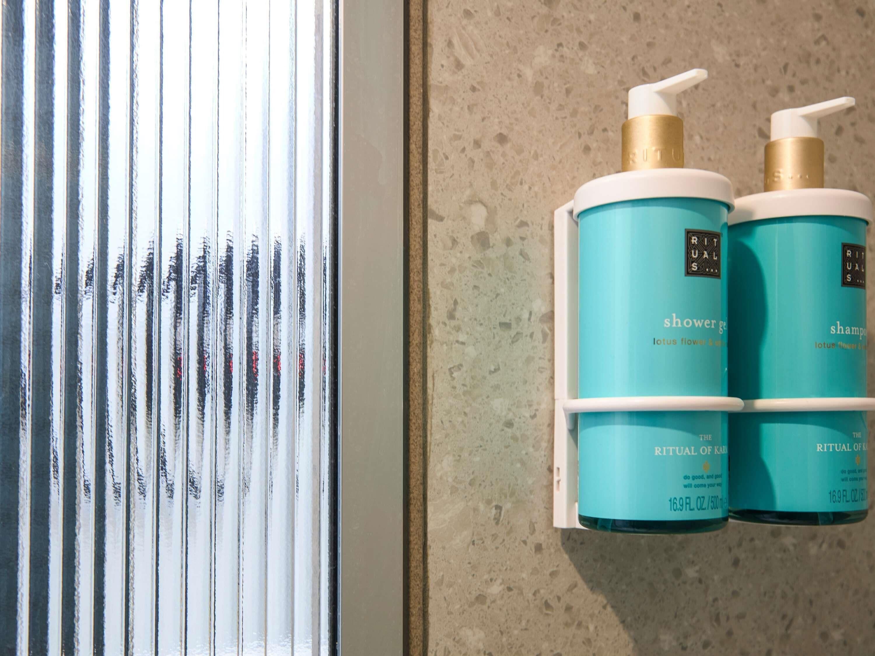 A bathroom with a window, a towel rack with two bottles of Rituals of Karma shower gel and shampoo, and a textured wall.