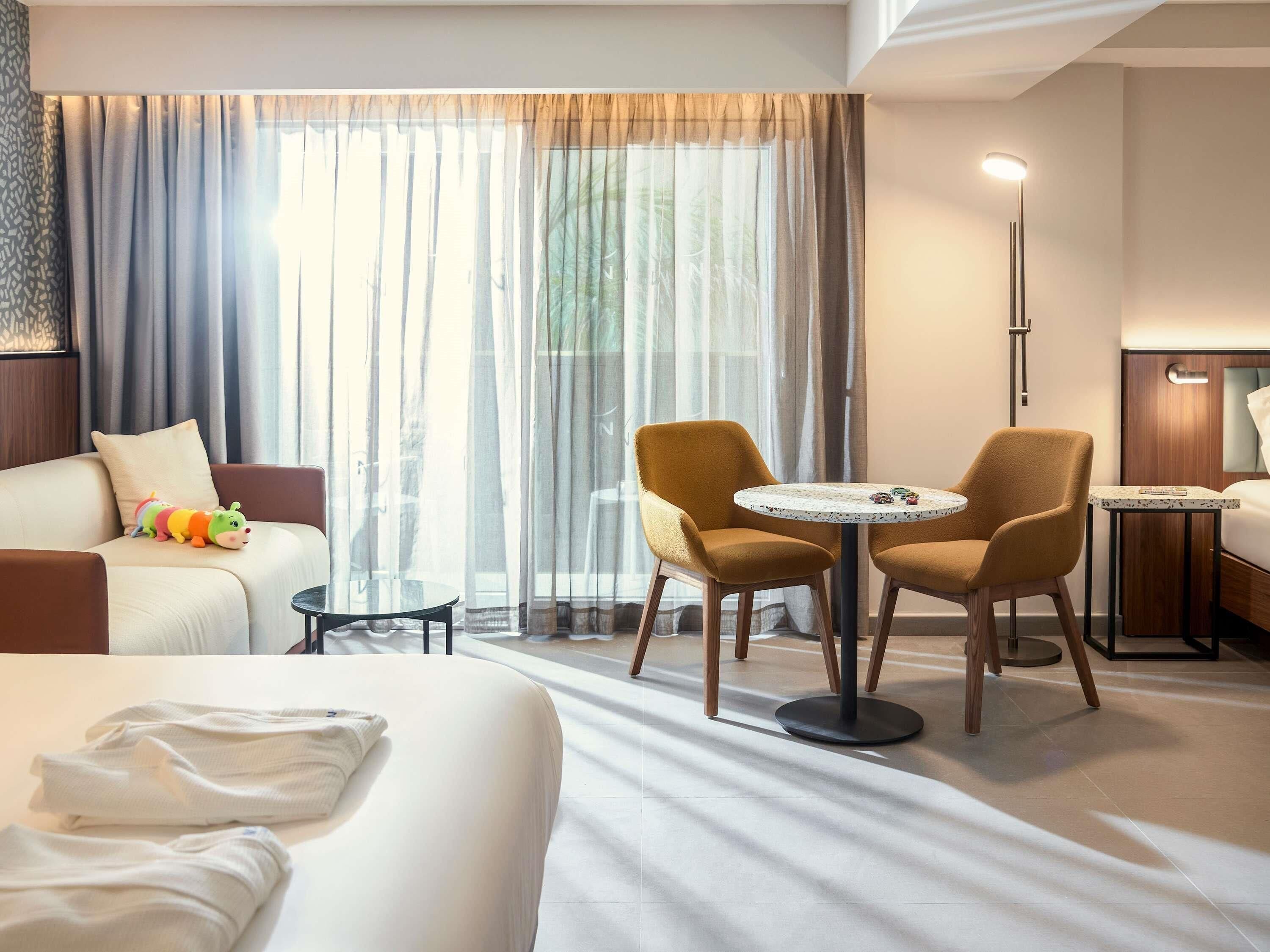 A hotel room with a bed, a sofa, two armchairs, a small round table, and a floor lamp.