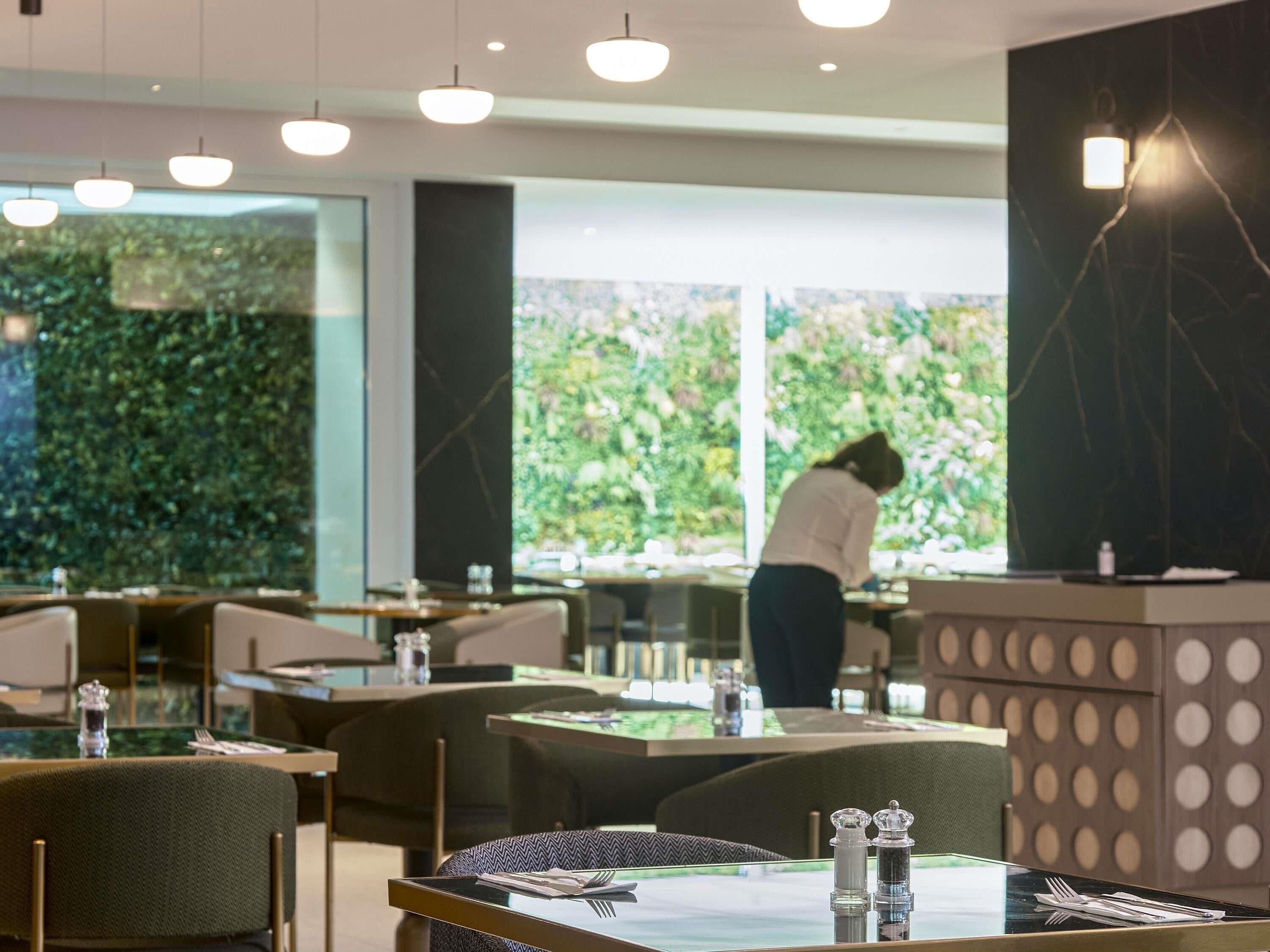 Un interno ristorante moderno con tavoli in vetro, sedie imbottite verdi e vista su un rigoglioso giardino attraverso ampie finestre.