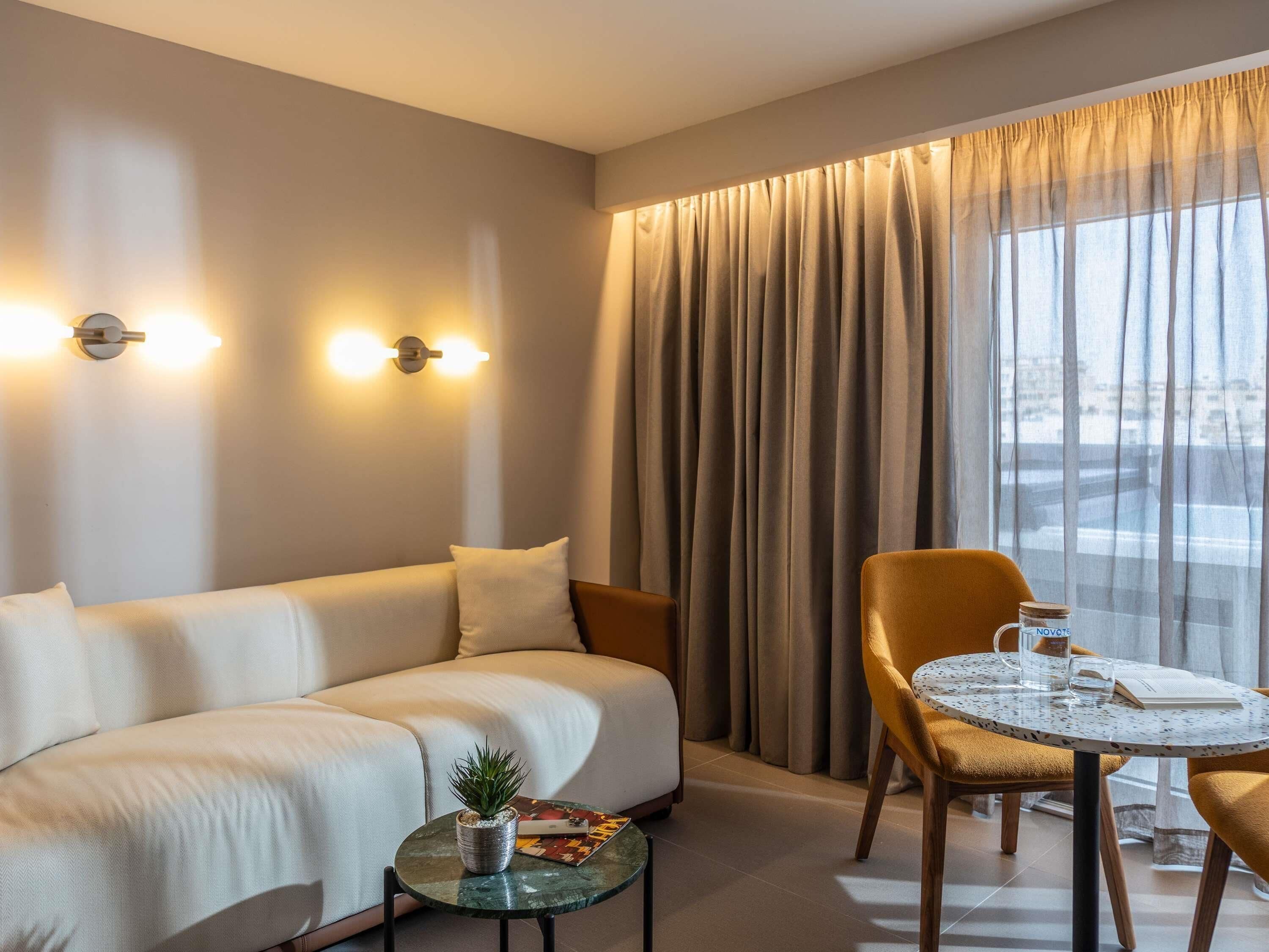 A modern hotel room with a sofa, a round table, a chair, a small potted plant, and a city view.