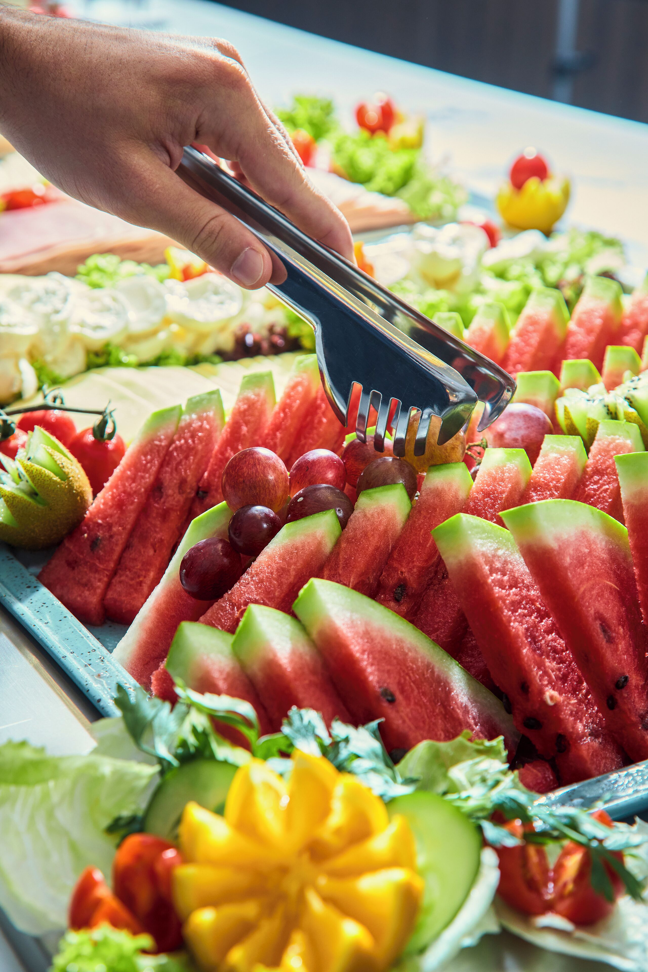 A hand using tongs to pick up a slice of watermelon from a tray of assorted fruits and vegetables.