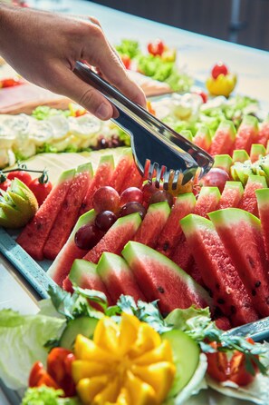 A hand using tongs to pick up a slice of watermelon from a tray of assorted fruits and vegetables. - Novotel Malta Sliema (Gzira)