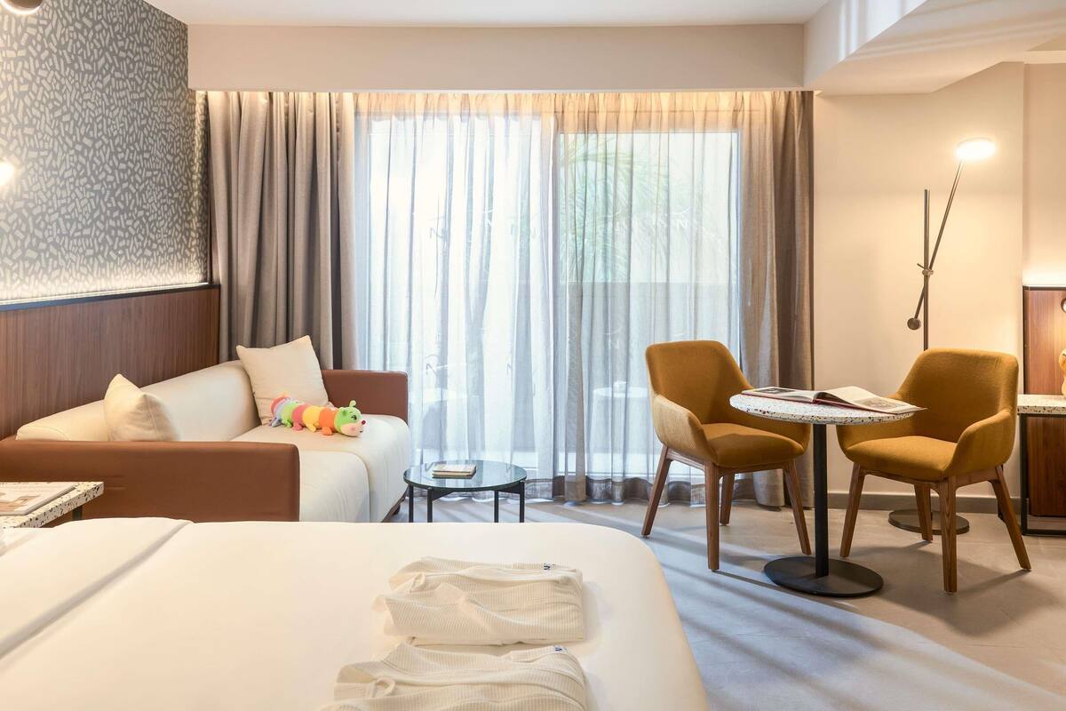 A hotel room with a bed, a sofa, a small table, two chairs, a lamp, and a window with curtains.