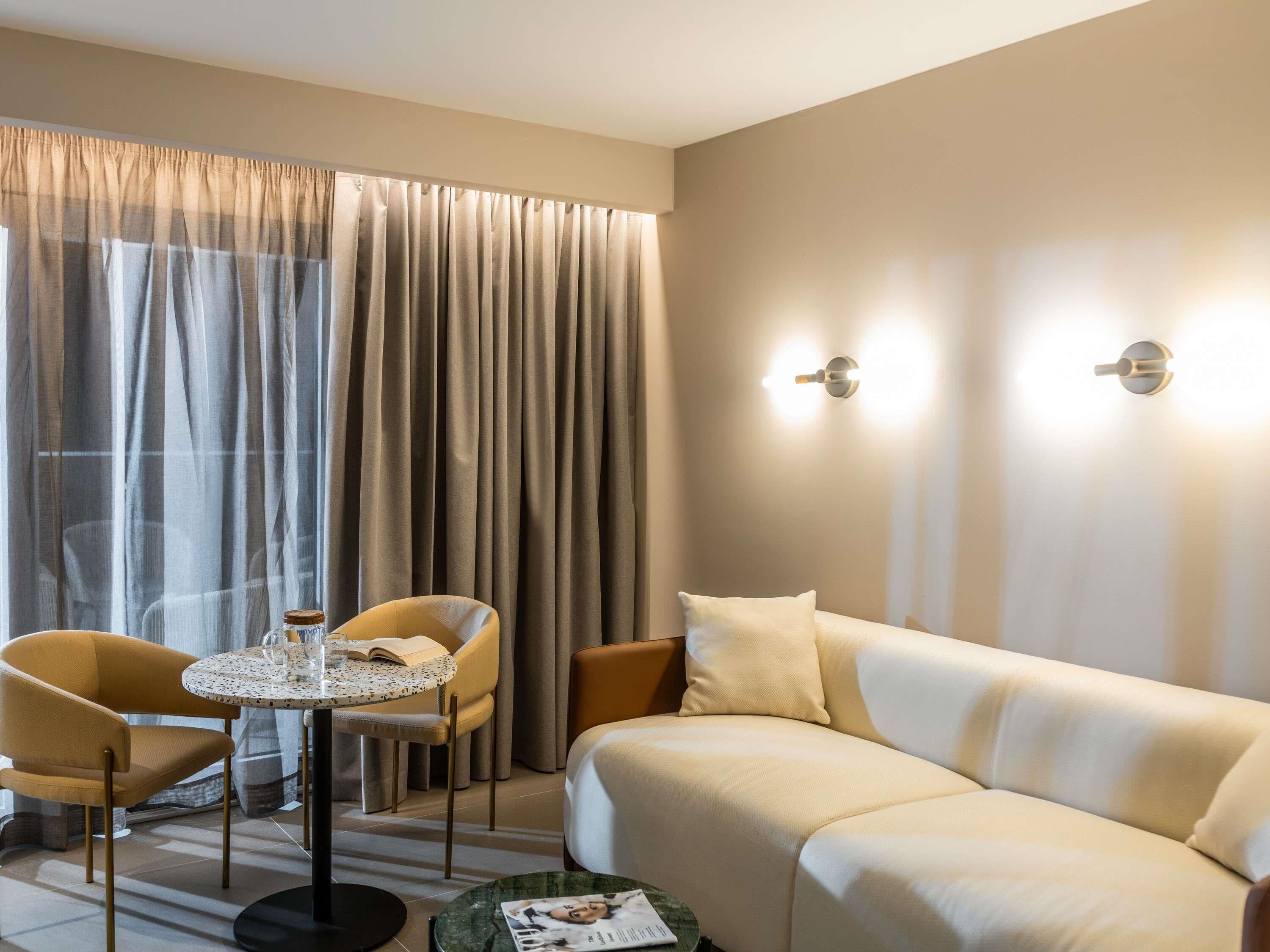 Une chambre d’hôtel moderne avec un canapé beige, deux fauteuils, une petite table ronde sur laquelle se trouvent un livre et un verre, et une grande fenêtre ornée de rideaux légers.