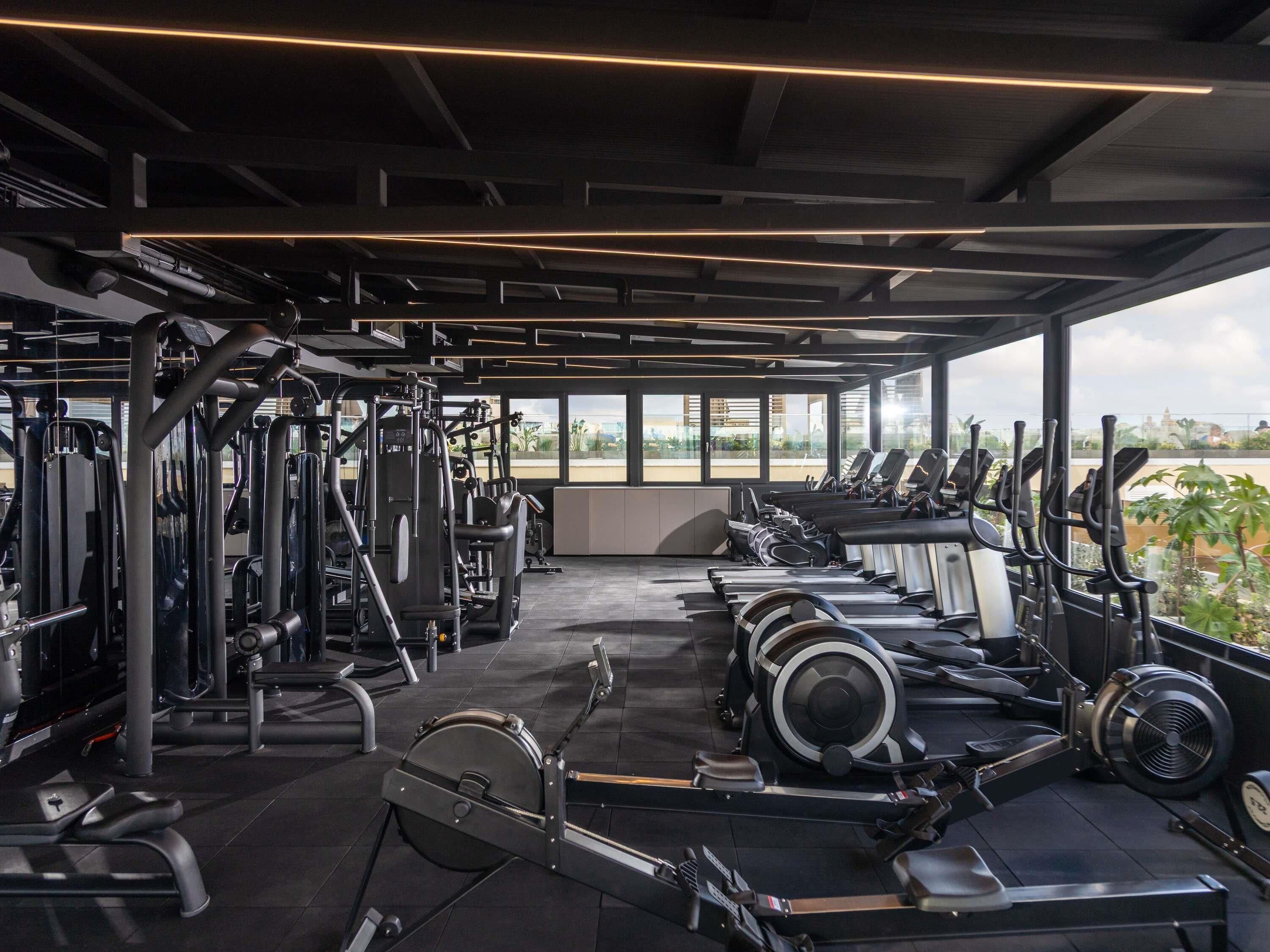 A gym with various exercise equipment, including treadmills and weight machines, under a covered area with large windows.