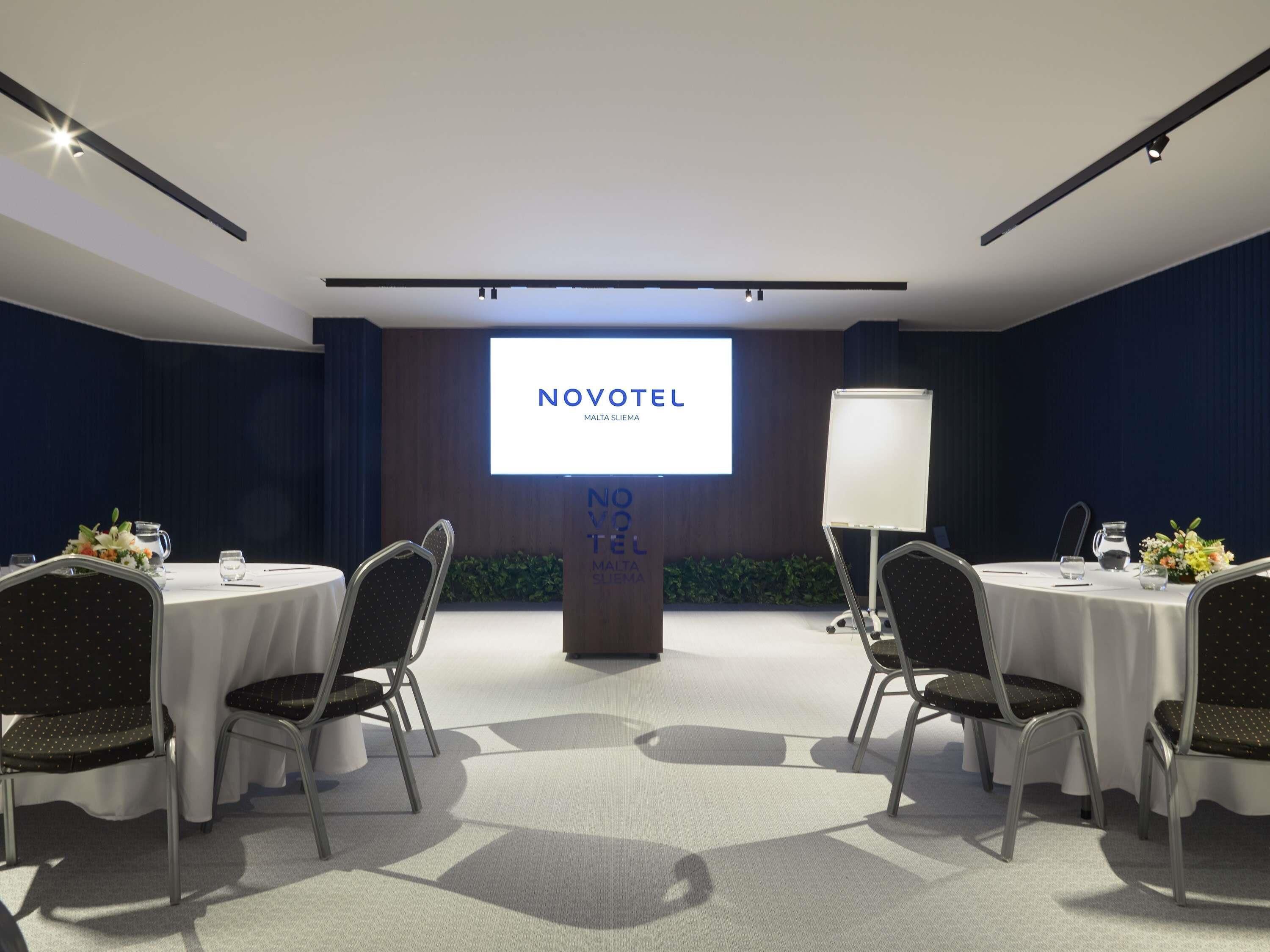 Une salle de conférence dotée d’un grand écran affichant le texte « NOVOTEL MALTE SLIEMA », d’un tableau blanc et de tables rondes entourées de chaises.