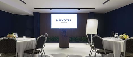 A conference room with a large screen displaying 'NOVOTEL MALTA SLIEMA', a whiteboard, and round tables with chairs.