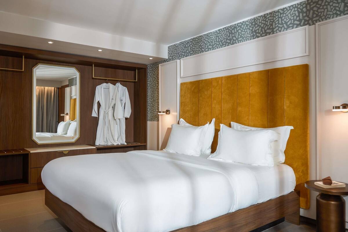 A modern hotel room with a large bed, a wooden headboard, a mirror, and a bedside table.
