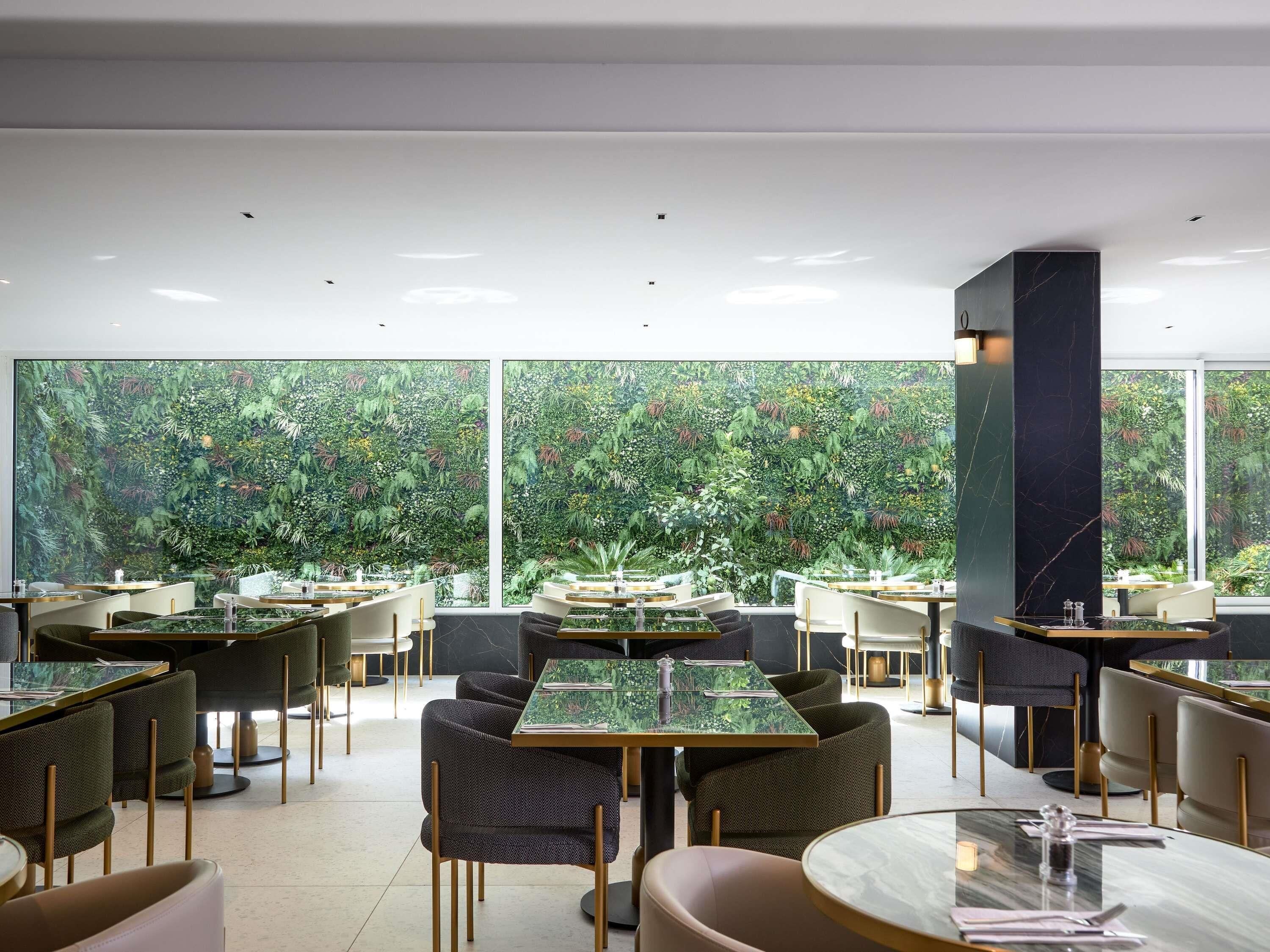 A modern restaurant with round tables and chairs, large windows with a view of greenery, and a marble column.