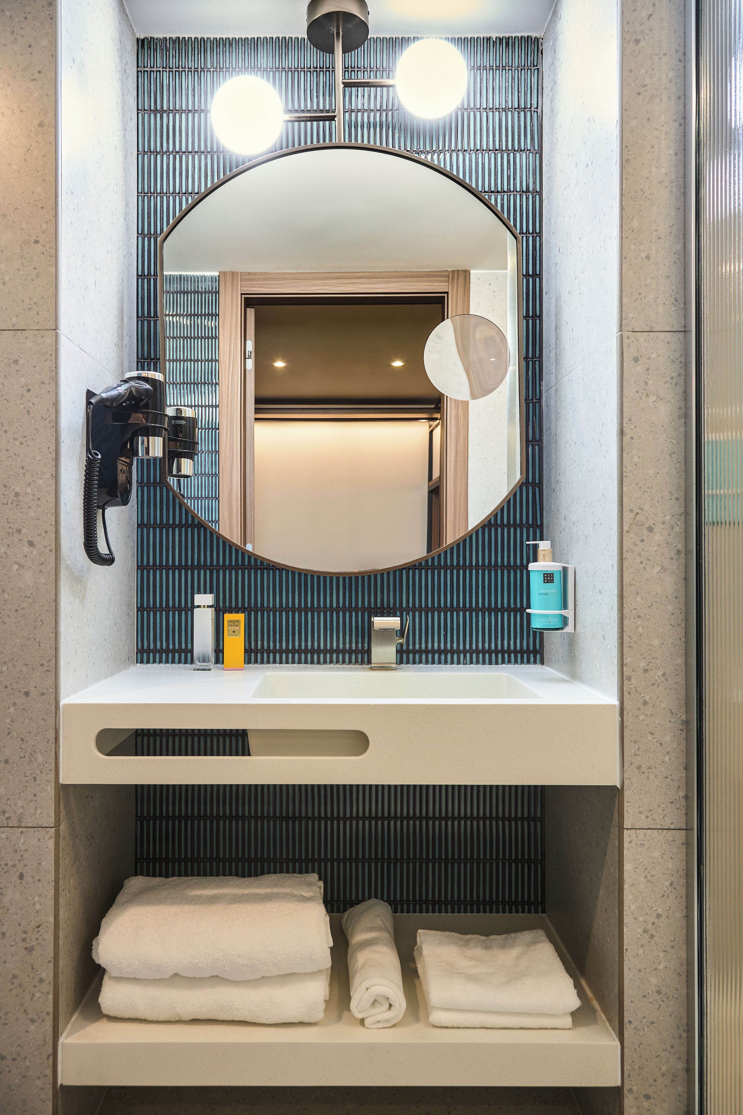 Une salle de bain moderne avec un miroir rond, un sèche-cheveux et des serviettes soigneusement pliées sur une étagère.