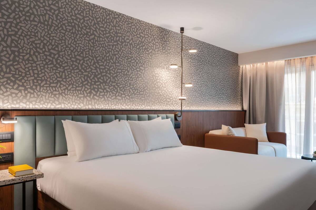 A modern hotel room with a large bed, a bedside table with a book, a nightstand, a lamp, and a patterned wall.