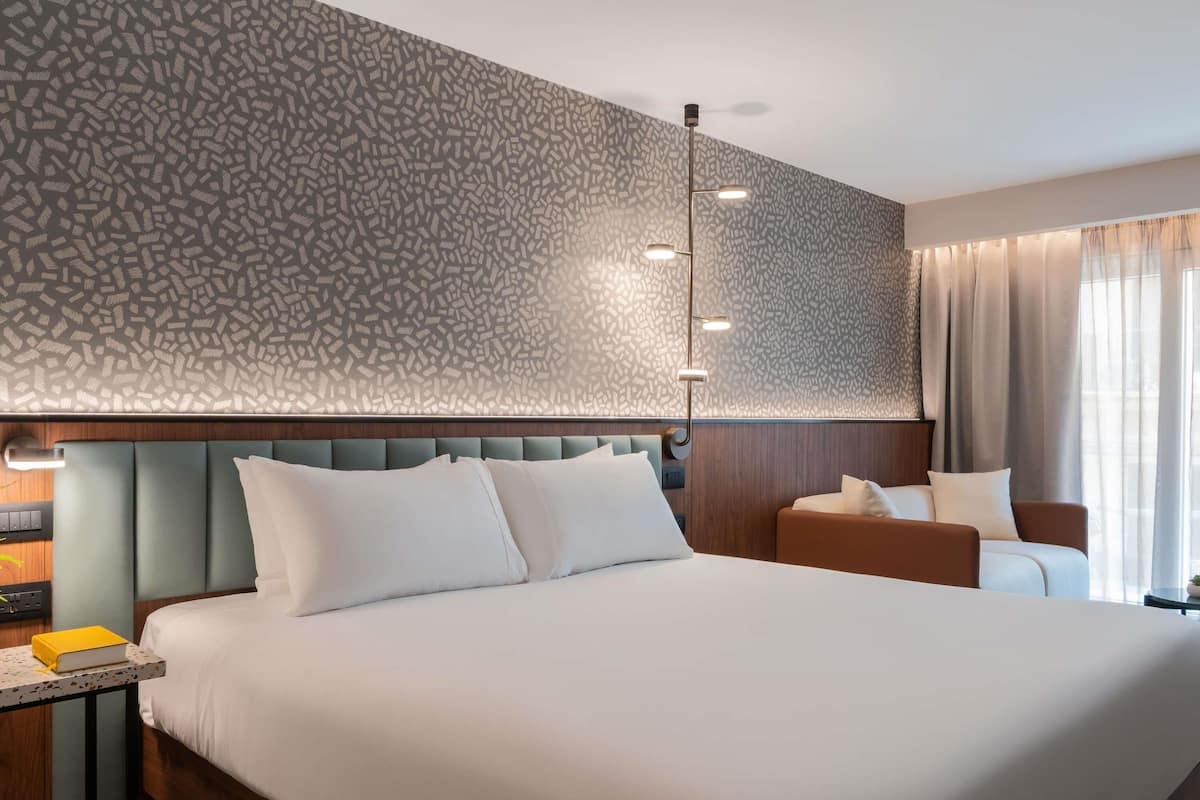 A modern hotel room with a large bed, a bedside table with a book, a nightstand, a lamp, and a patterned wall.