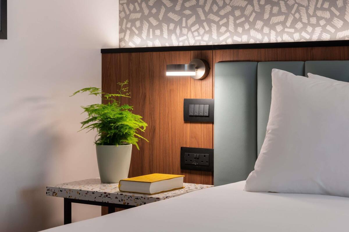 A hotel room with a bed, a bedside table with a book and a plant, a wall-mounted light, and a power outlet.