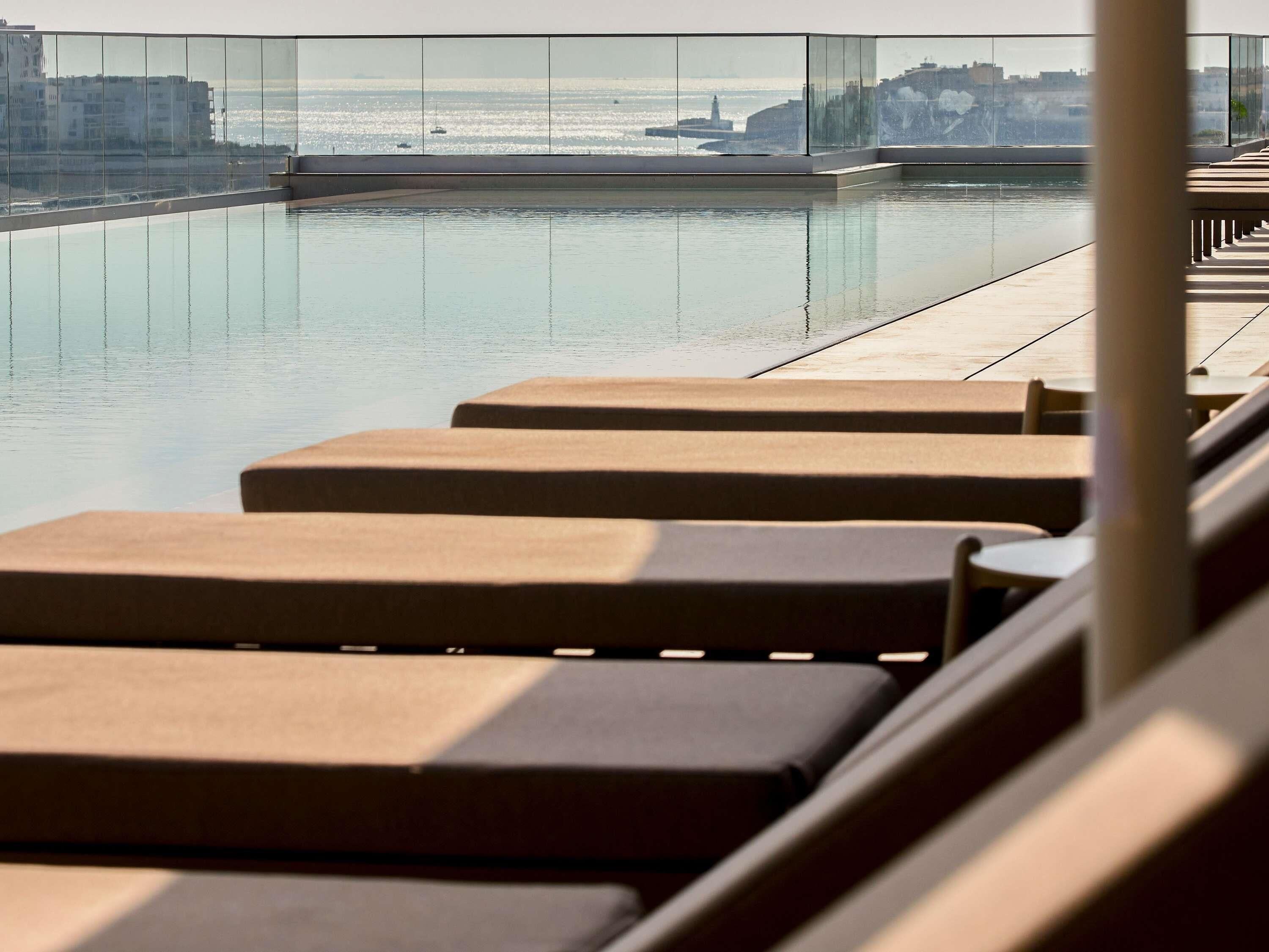 A rooftop pool with lounge chairs and a glass balustrade overlooking the sea.