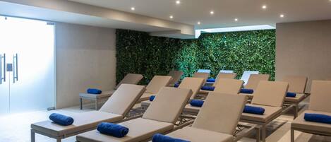 A modern spa room with multiple sun loungers, a green wall, and recessed ceiling lights.