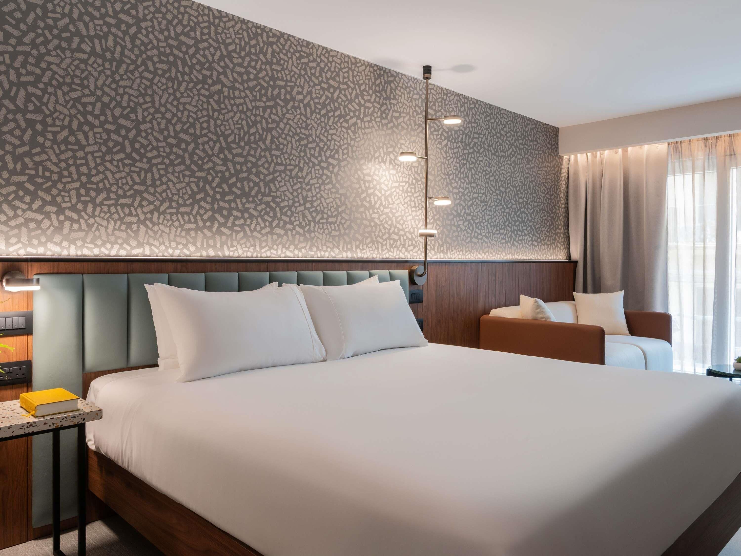 A hotel room with a large bed, a bedside table with a book, a nightstand, a wall-mounted lamp, and a window with curtains.