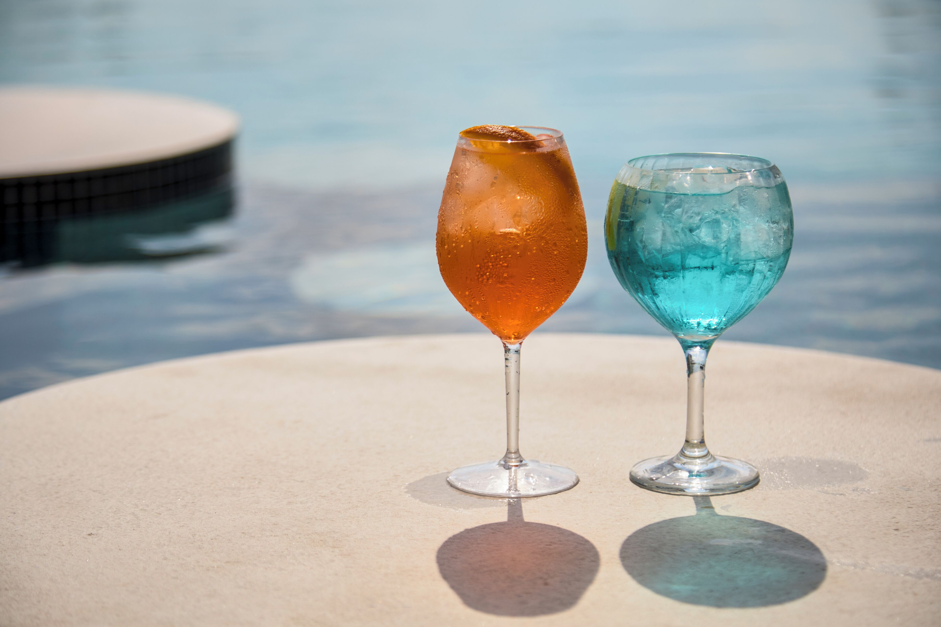 Two glasses of beverages on a table by a pool.