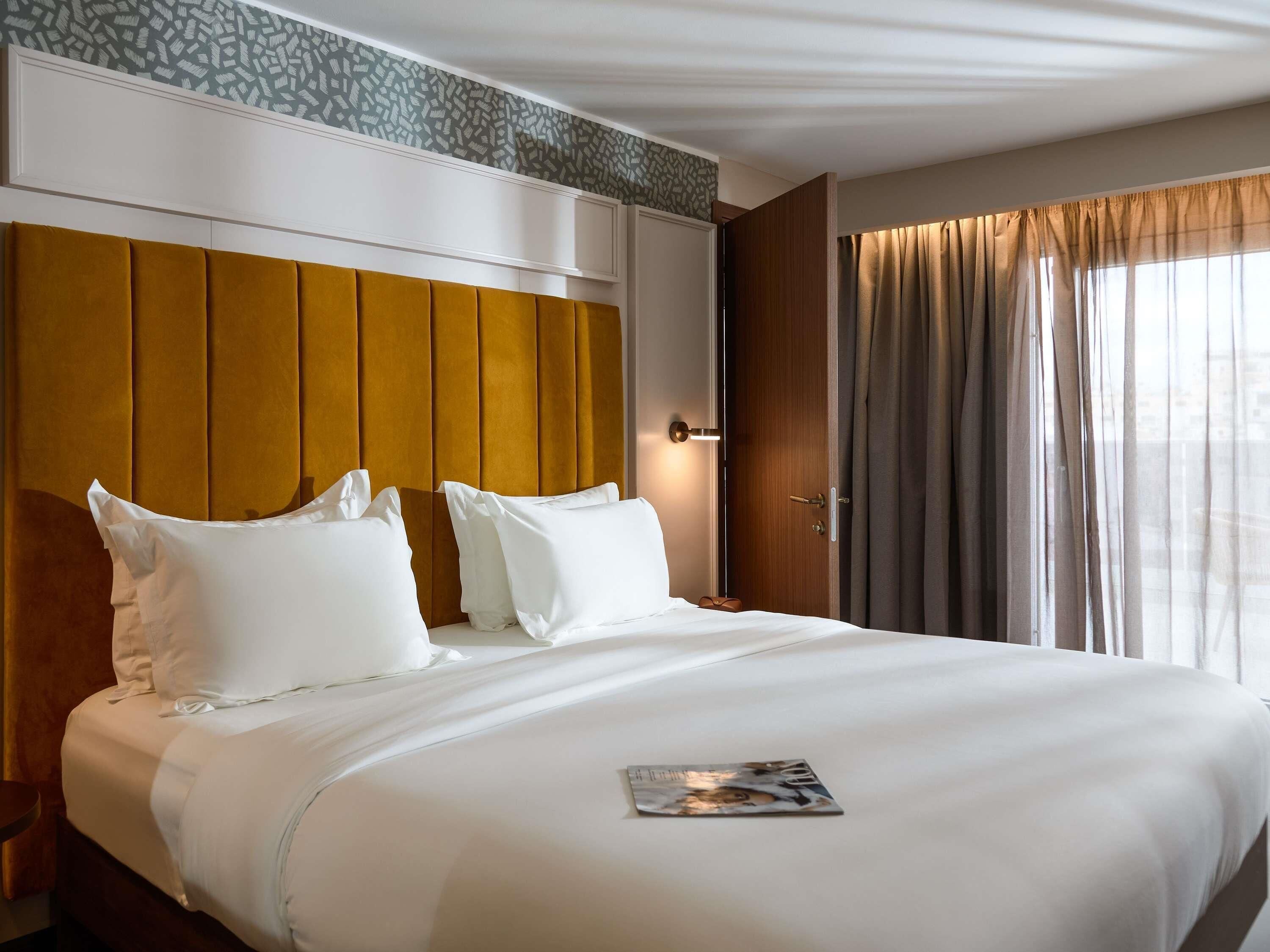 A hotel room with a large bed, white bedding, a magazine on the bed, a wooden headboard, and a window with curtains.