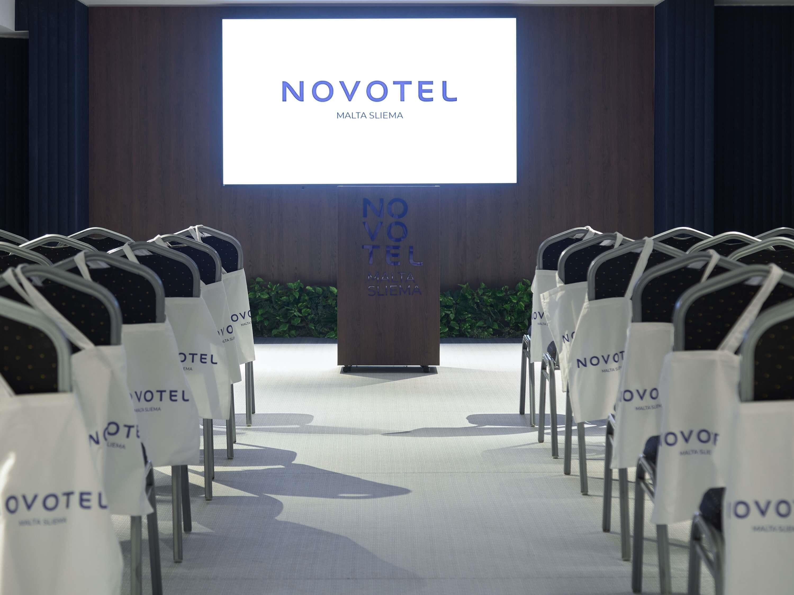 Una sala de conferencias con filas de sillas frente a un atril con una pantalla que muestra 'NOVOTEL MALTA SLIEMA'.