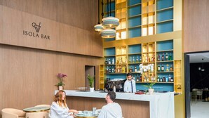 A modern bar with a wooden wall and a sign reading "ISOLA BAR". There are two people seated at a table, and a bartender is behind the counter. - Novotel Malta Sliema (Gzira)