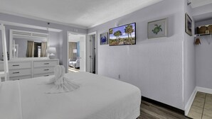 Habitación de hotel con una cama grande, un aparador, un espejo y una puerta que da a otra habitación. Hay dos cuadros en la pared.
