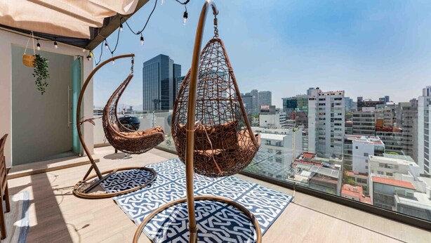 A rooftop terrace with hanging chairs, patterned rugs, and a city view.