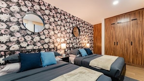 Une chambre avec deux lits, un papier peint à motifs floraux, une armoire en bois et un miroir rond.
