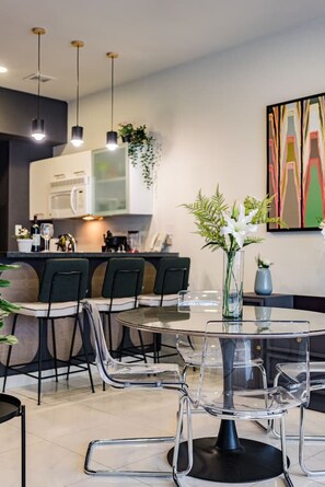 Een moderne eetruimte met een glazen tafel en stoelen, een keuken op de achtergrond en een decoratieve plant aan de muur.