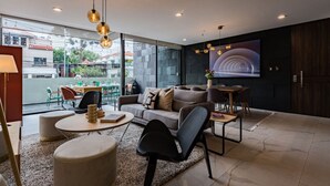 Un salon moderne comprenant un canapé, une table basse et des fauteuils, un téléviseur et une grande fenêtre donnant sur des immeubles.