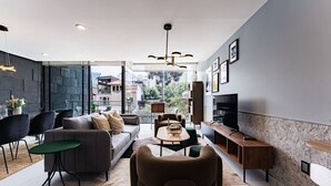 Una sala de estar moderna con un sofá, sillas, una mesa de centro y un televisor. Hay un ventanal que ofrece vistas de la ciudad.
