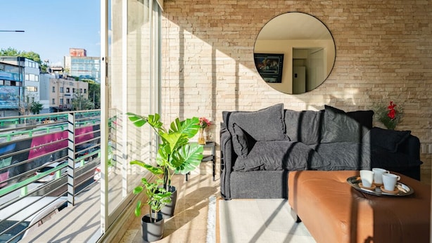 Un balcone con vista sulla città, uno specchio rotondo al muro, un divano grigio e piante in vaso.