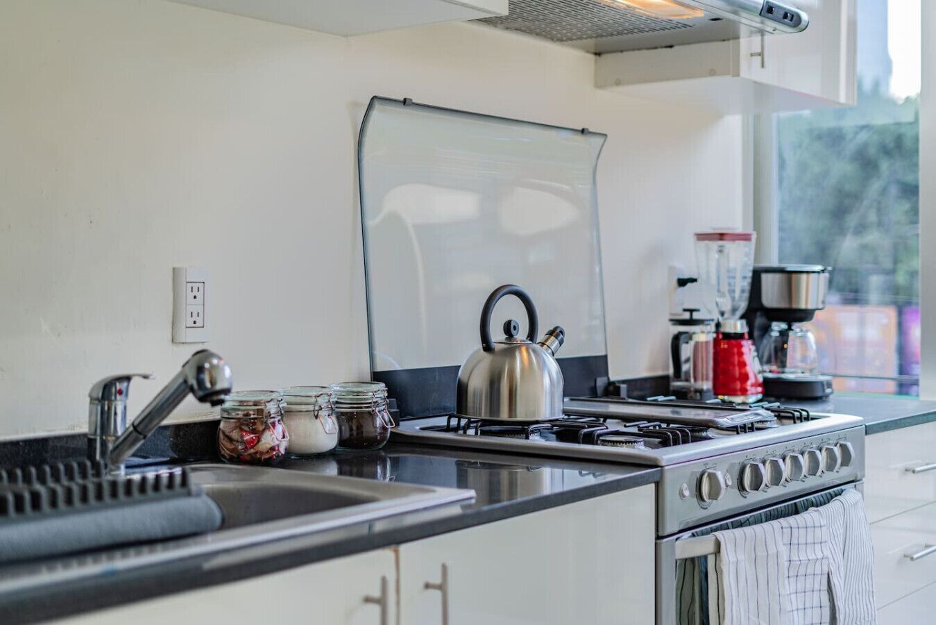 A modern kitchen with a gas stove, kettle, and coffee maker. There are jars of spices on the counter, and a window provides natural light.