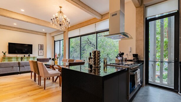 A modern kitchen with a large island, a dining area with a sofa and chairs, and a large window with a view of trees.
