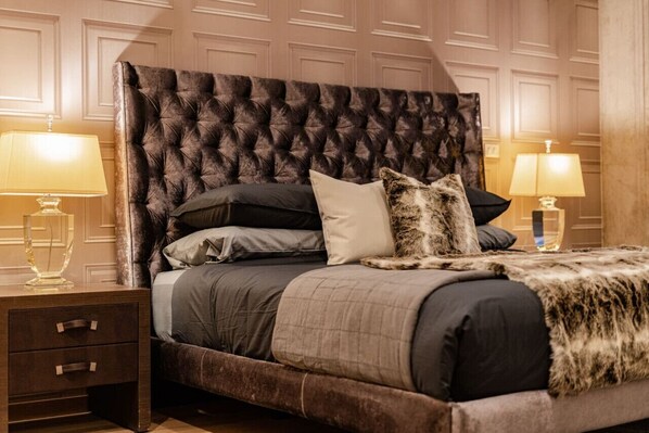 A bedroom with a tufted headboard, a bed with grey bedding, two bedside tables with lamps, and a nightstand with drawers.