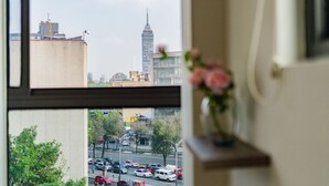 A window view of a cityscape with a tall building, a busy street, and a vase with flowers on a shelf.