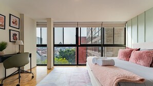 A modern bedroom with a large bed, a desk, and a view of the city through floor-to-ceiling windows.