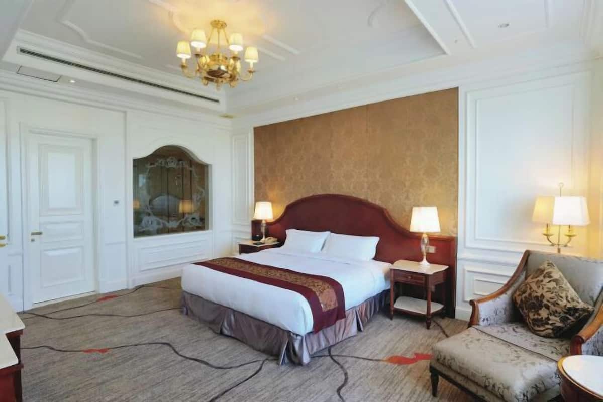 Hotelkamer met een groot bed, twee nachtkastjes met lampen, een rood bekleed hoofdeinde, een grijze fauteuil en een geborduurde plaid.
