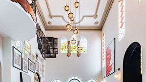Un vaste salon doté d’un haut plafond, d’un canapé en cuir noir, d’un fauteuil noir et d’une table basse. Les murs sont ornés de tableaux encadrés et un lustre est suspendu au plafond.