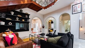 Un salon moderne avec un canapé en cuir marron, des fauteuils noirs et une table basse. On y trouve des étagères ornées d’objets décoratifs et un grand miroir. Des portes en arc donnent accès aux autres pièces.