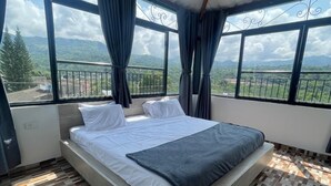 Ein Schlafzimmer mit einem großen Bett, zwei Fenstern mit Vorhängen und Blick auf eine gebirgige Landschaft.