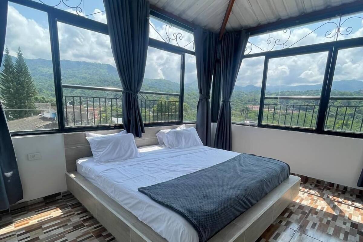 Une chambre à coucher avec un grand lit, deux fenêtres avec des rideaux et une vue sur un paysage montagneux.