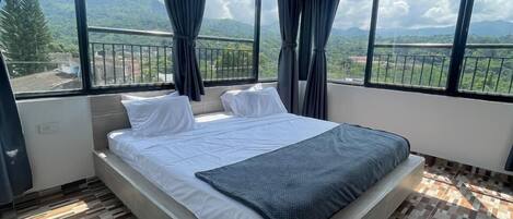 A bedroom with a large bed, two windows with curtains, and a view of a mountainous landscape.