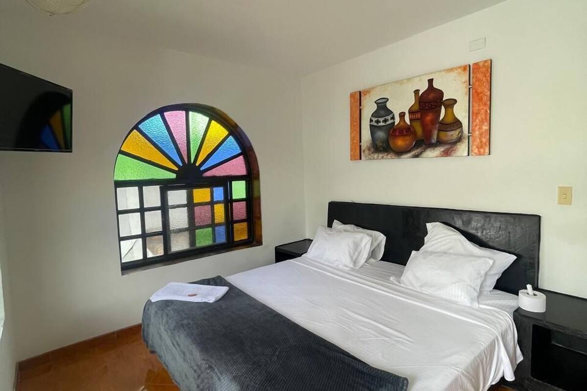 Une chambre à coucher comprenant un lit, une table de chevet, un ventilateur fixé au mur, une fenêtre ornée de vitraux colorés et un tableau représentant des poteries.