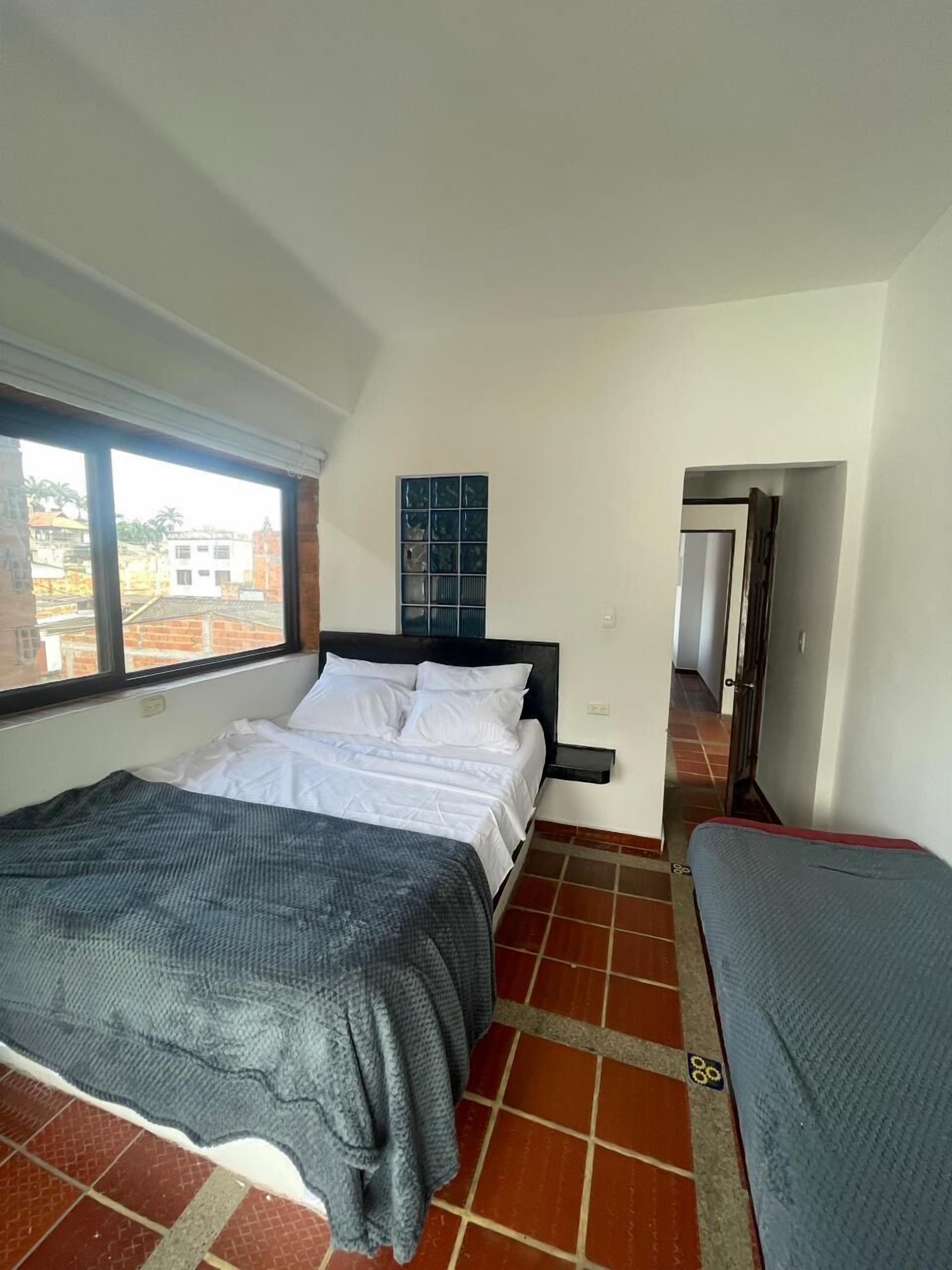 A bedroom with a bed, a window, a nightstand, and a view of buildings outside.