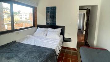 A bedroom with a bed, a window, a nightstand, and a view of buildings outside.