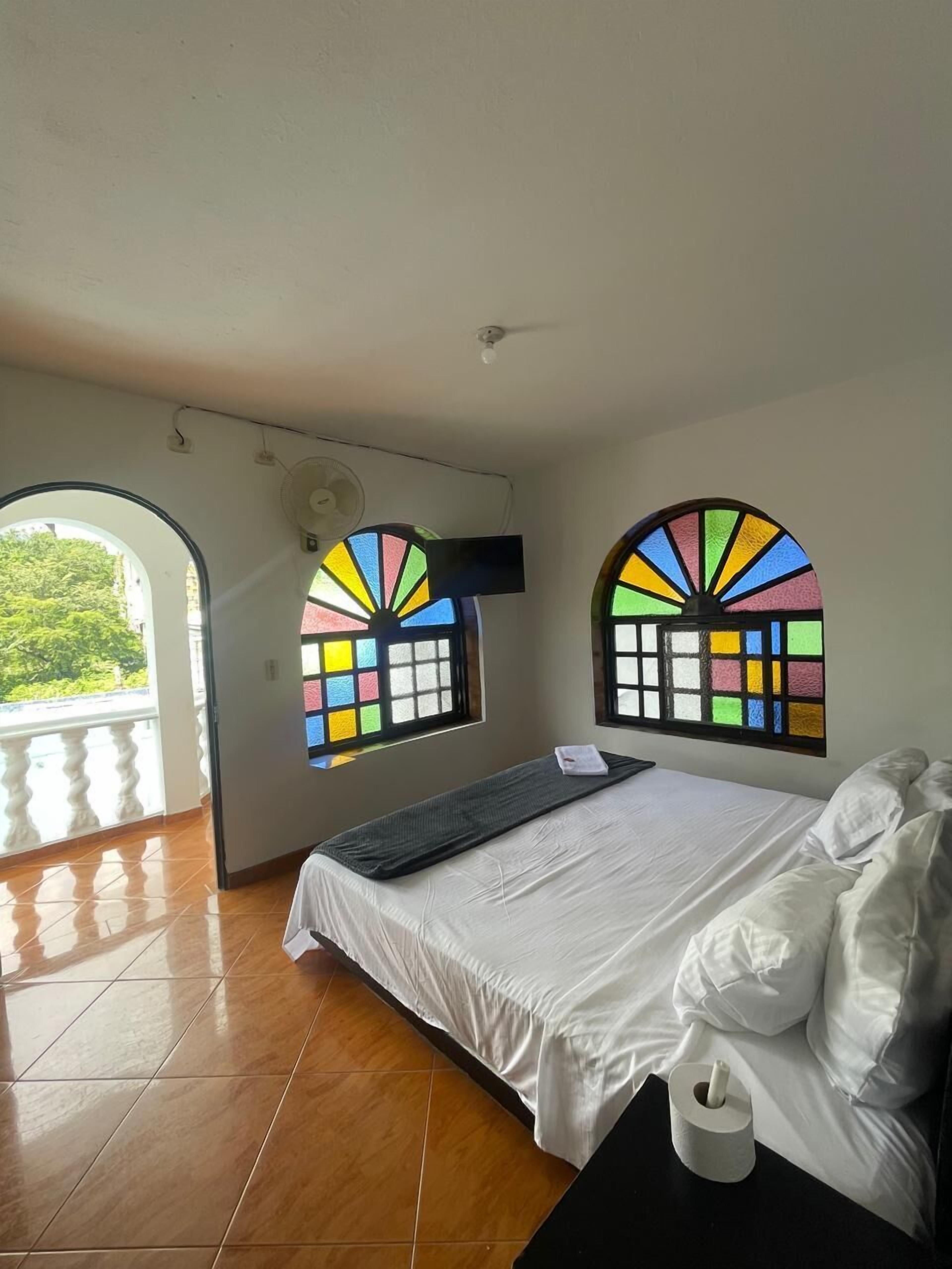 Un dormitorio con cama, ventana con vidrio de colores, ventilador de techo y balcón.
