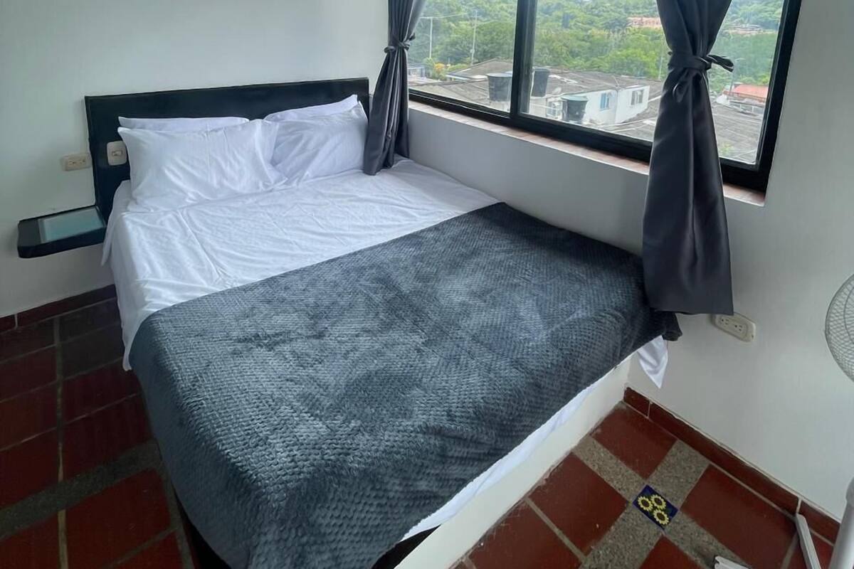 Un lit simple avec une couverture grise dans une chambre dotée d’une fenêtre donnant sur un paysage montagneux.