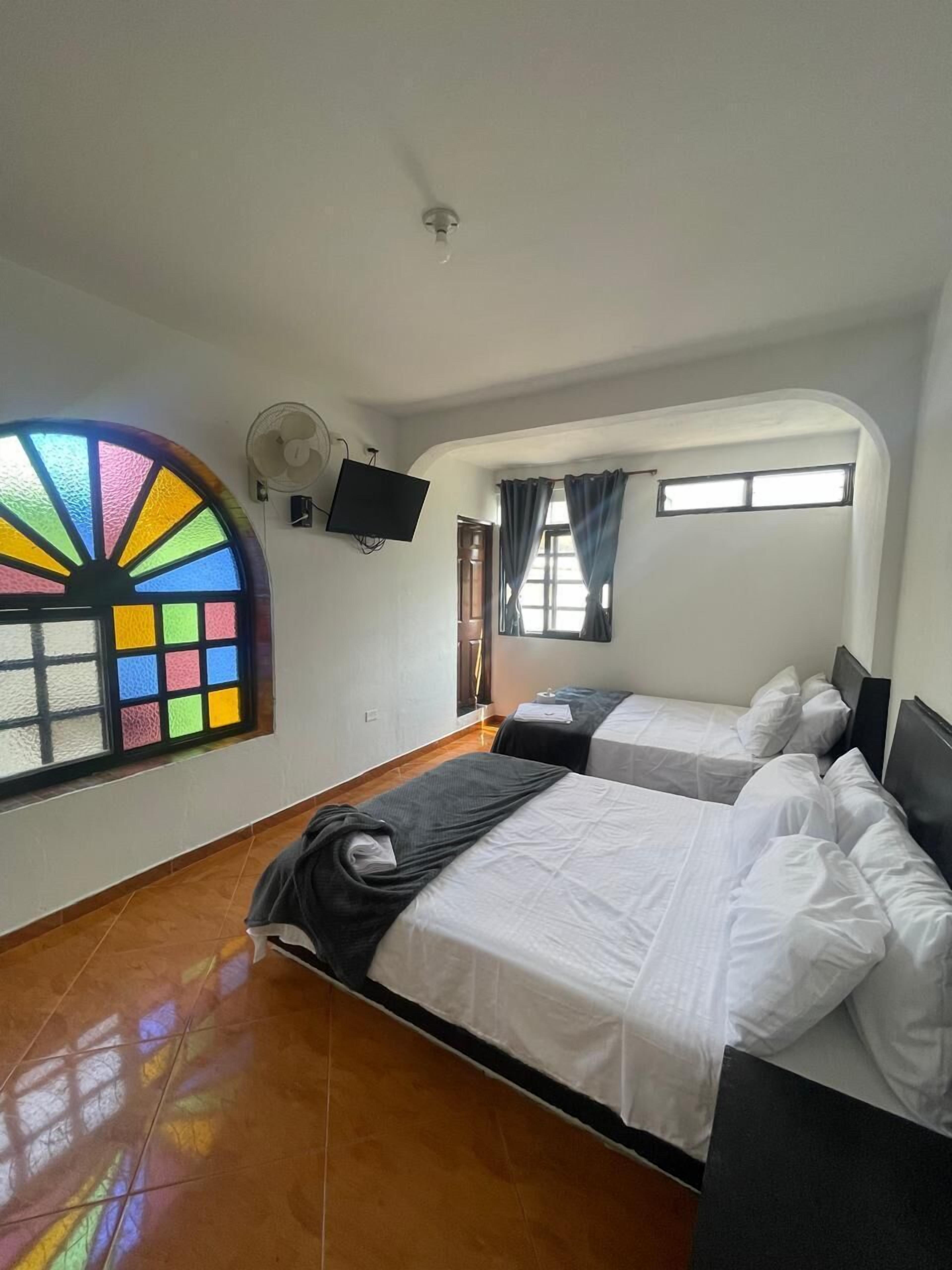 A hotel room with two beds, a large window with colorful stained glass, a ceiling fan, and a flat-screen TV mounted on the wall.